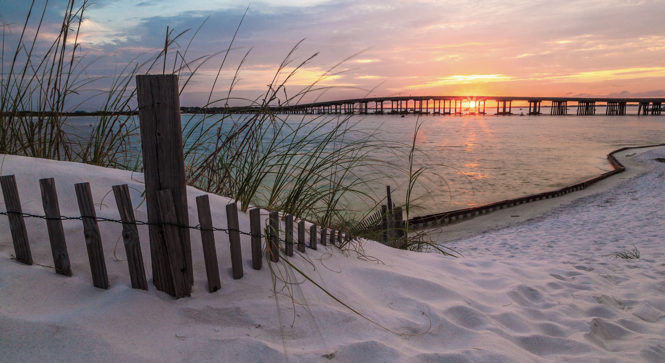Sunset over sandy beach with bridge silhouette.